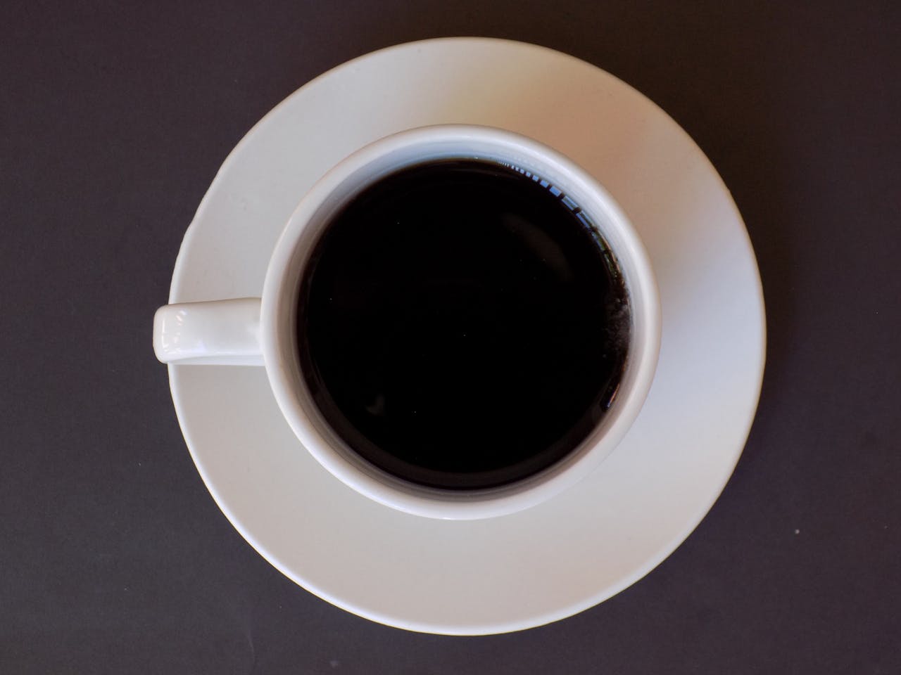 High-angle photo of black coffee in a white cup and saucer on a dark background, ideal for food and beverage themes.