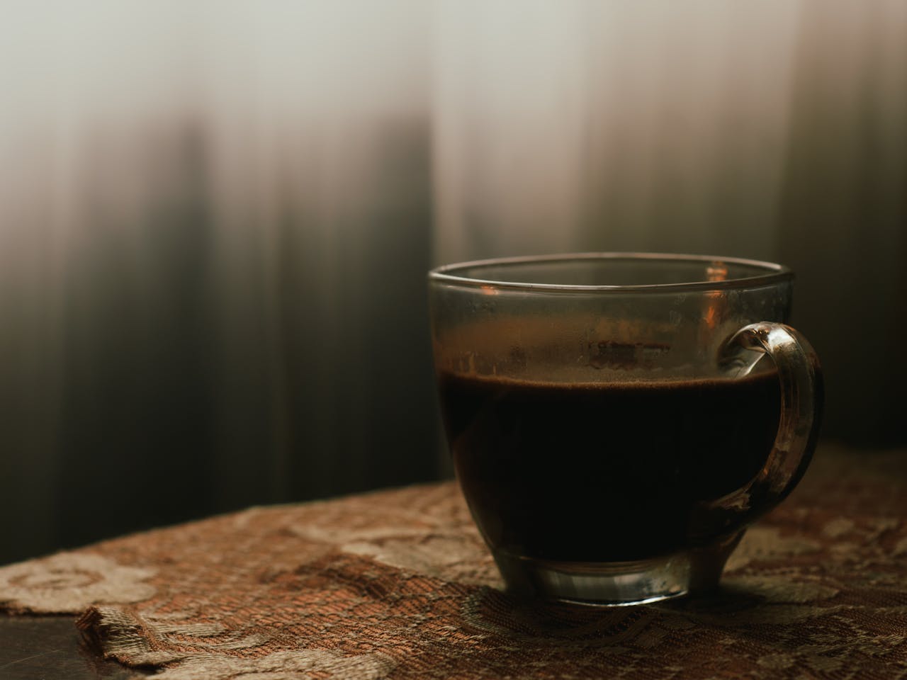 A tranquil scene featuring a glass cup of black coffee on a patterned cloth in soft window light.